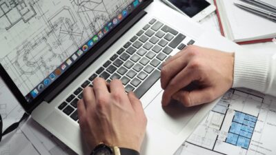 A person's hands typing on a laptop with architectural blueprints displayed on the screen and spread on the desk