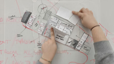 a woman's hands with a pencil over an architectural plan on a desk.