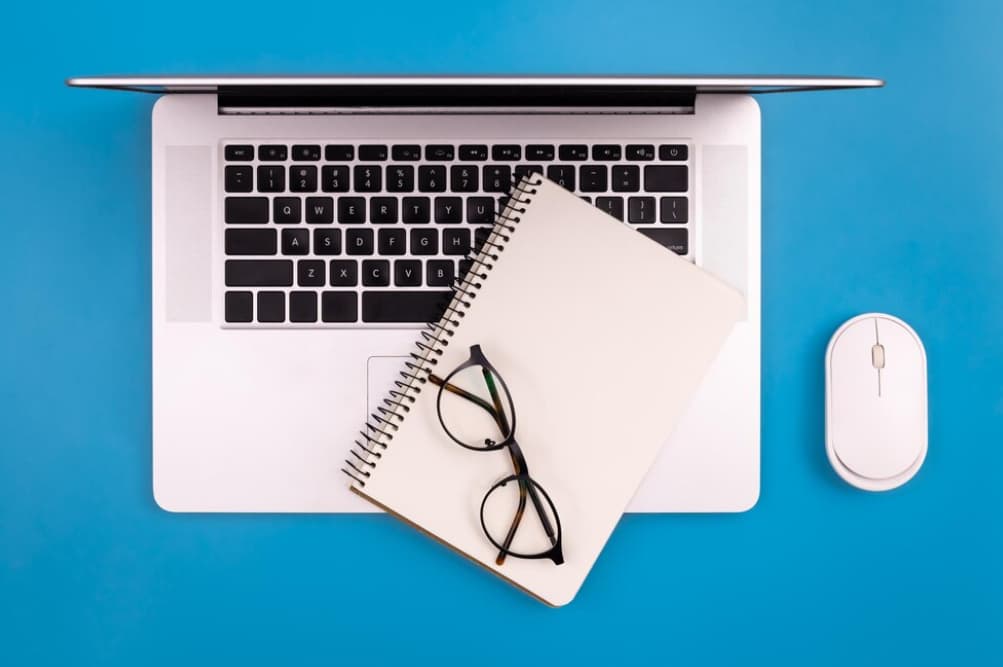 A neat workspace with an open laptop, notepad, glasses, and mouse on a blue background