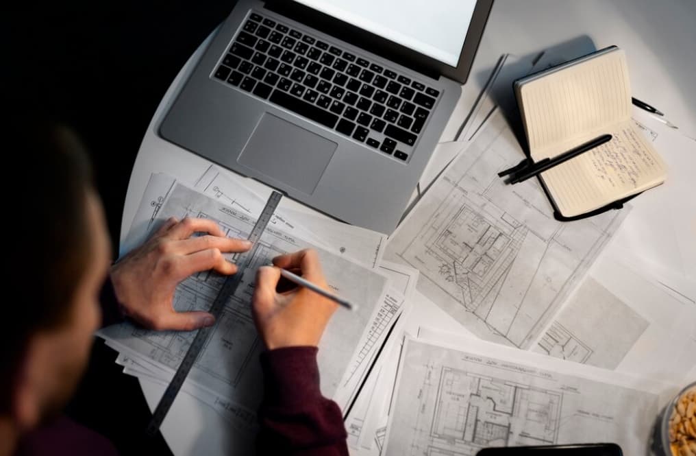 An architect working on blueprints next to a laptop and notebook